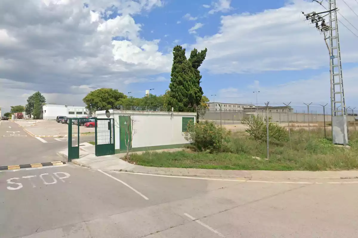 Una carretera con señal de stop y un portón verde al lado de un edificio cercado con alambre de púas y árboles en el fondo bajo un cielo parcialmente nublado.