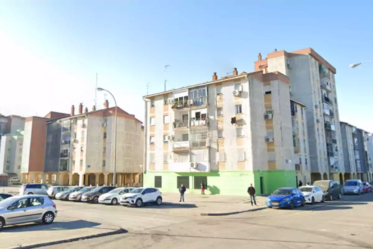 Edificios residenciales de varios pisos con coches aparcados en la calle y personas caminando en un día soleado.