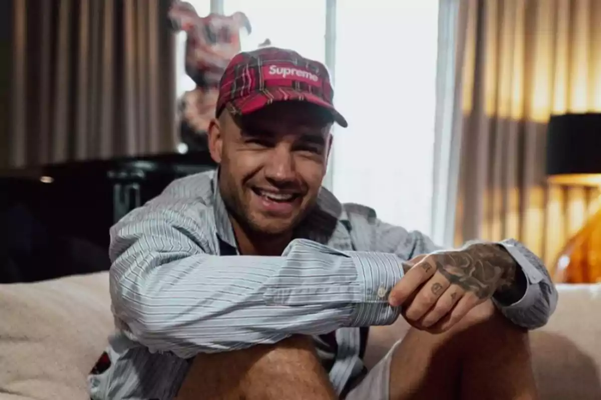 Un hombre sonriente con gorra roja y camisa a rayas sentado en un sofá.