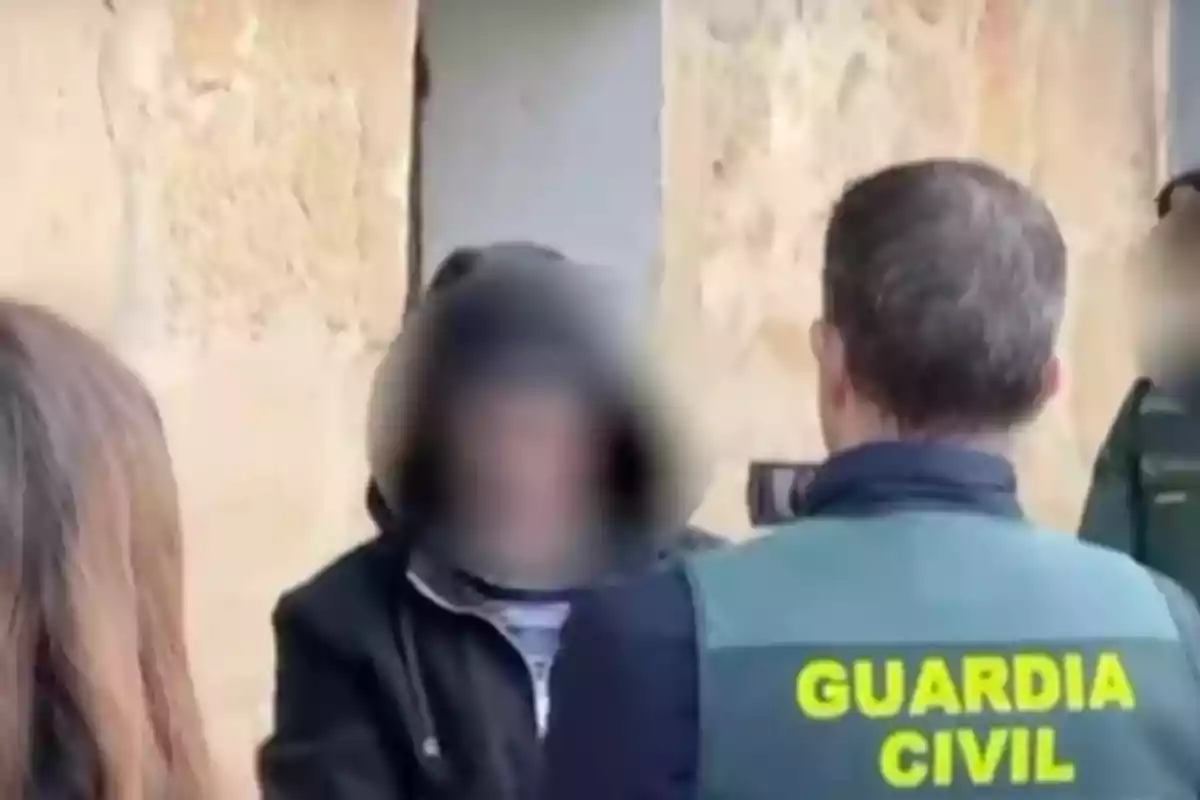 Un agente de la Guardia Civil interactúa con una persona con el rostro difuminado frente a una pared de piedra.