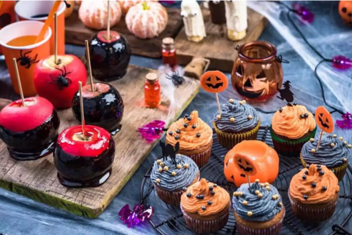 Mesa decorada con dulces temáticos de Halloween que incluye manzanas acarameladas y cupcakes con glaseado naranja y negro adornados con figuras de calabazas y brujas.