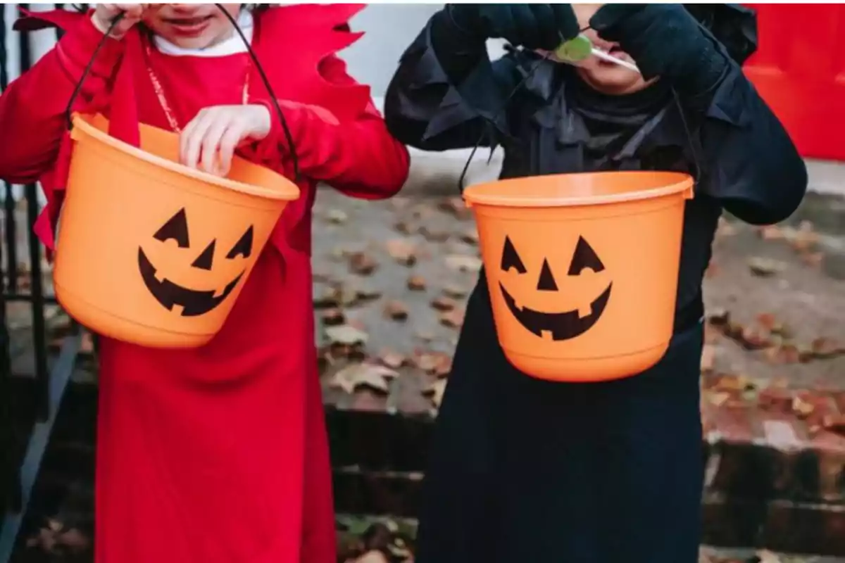 Dos niños disfrazados sostienen cubetas naranjas con caras de calabaza para recolectar dulces en Halloween.