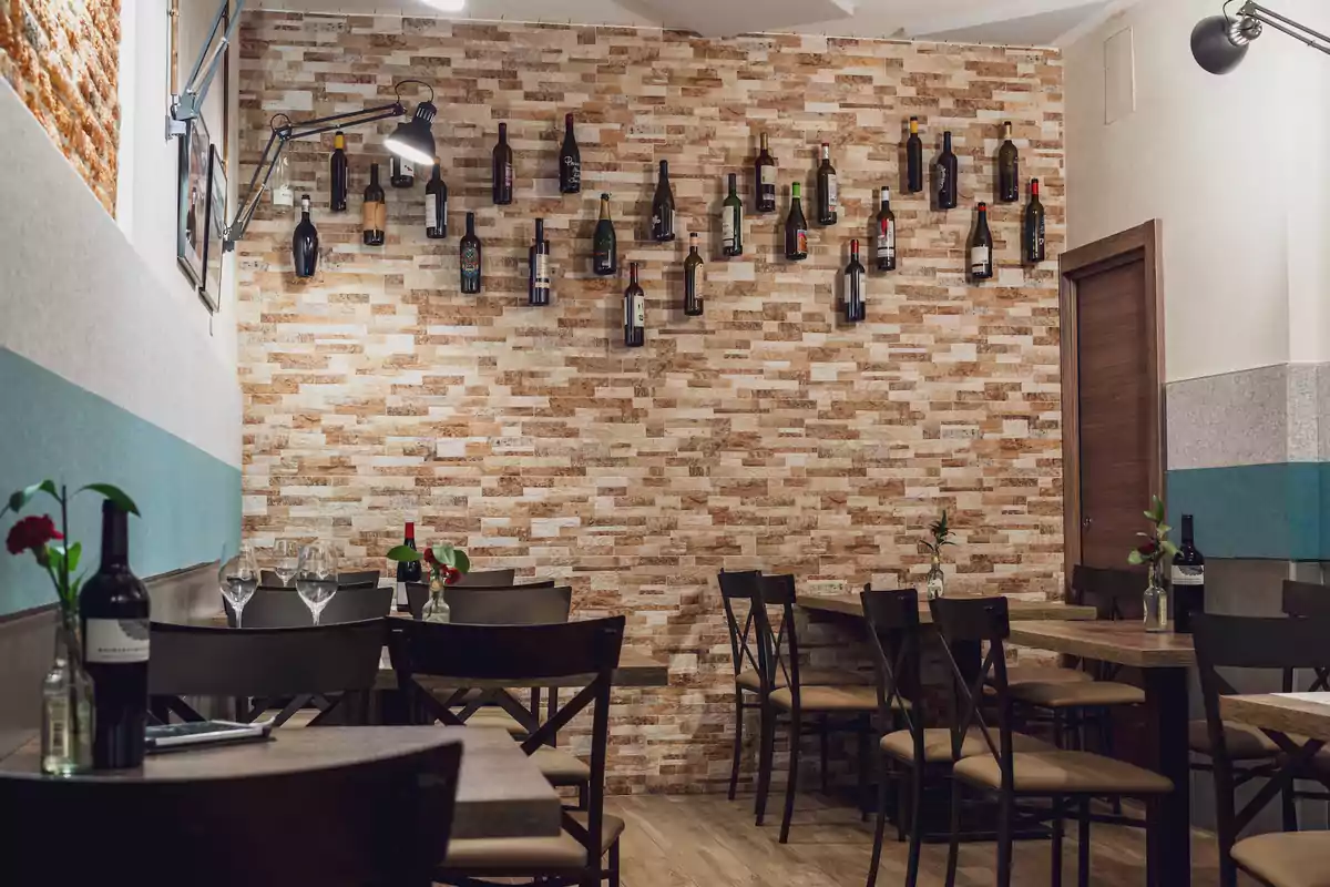 Un acogedor restaurante con mesas de madera y sillas negras, decorado con botellas de vino en una pared de ladrillo.