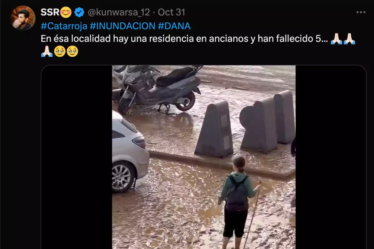 Una persona camina por una calle inundada y llena de barro, con vehículos y contenedores al fondo.