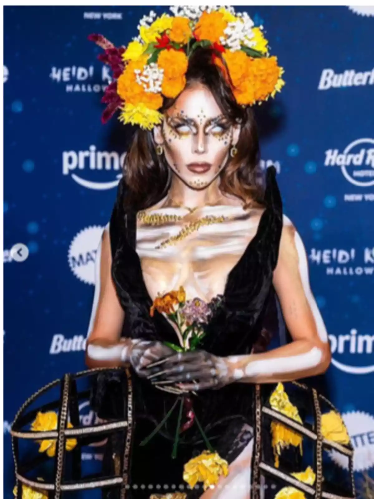 Persona con maquillaje artístico y corona de flores, sosteniendo un ramo, en un evento con fondo de logotipos.