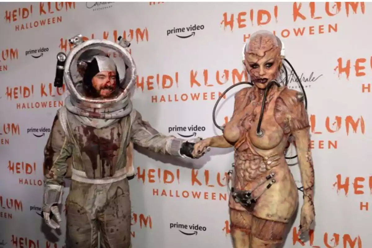 Dos personas disfrazadas de astronauta y criatura alienígena posan en un evento de Halloween con un fondo que dice 