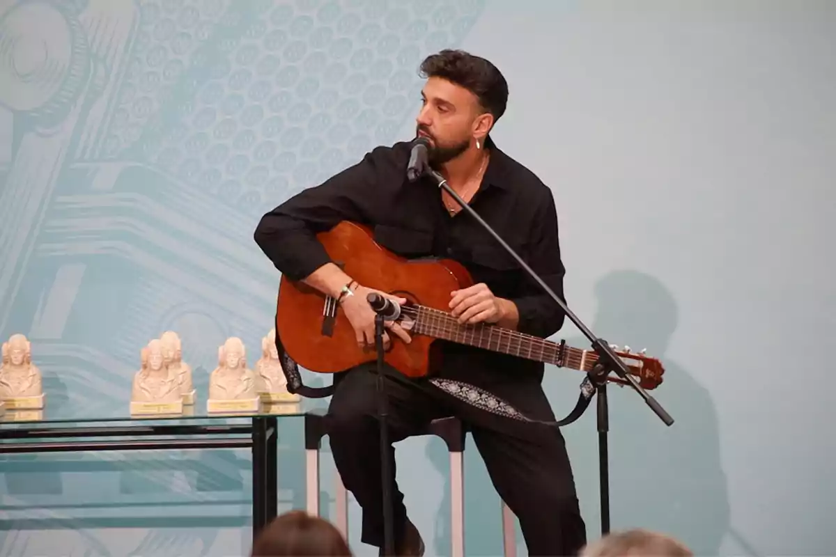 Carlos Peralías  tocando la guitarra y cantando en el centenario de Radio España con varias estatuillas sobre una mesa.