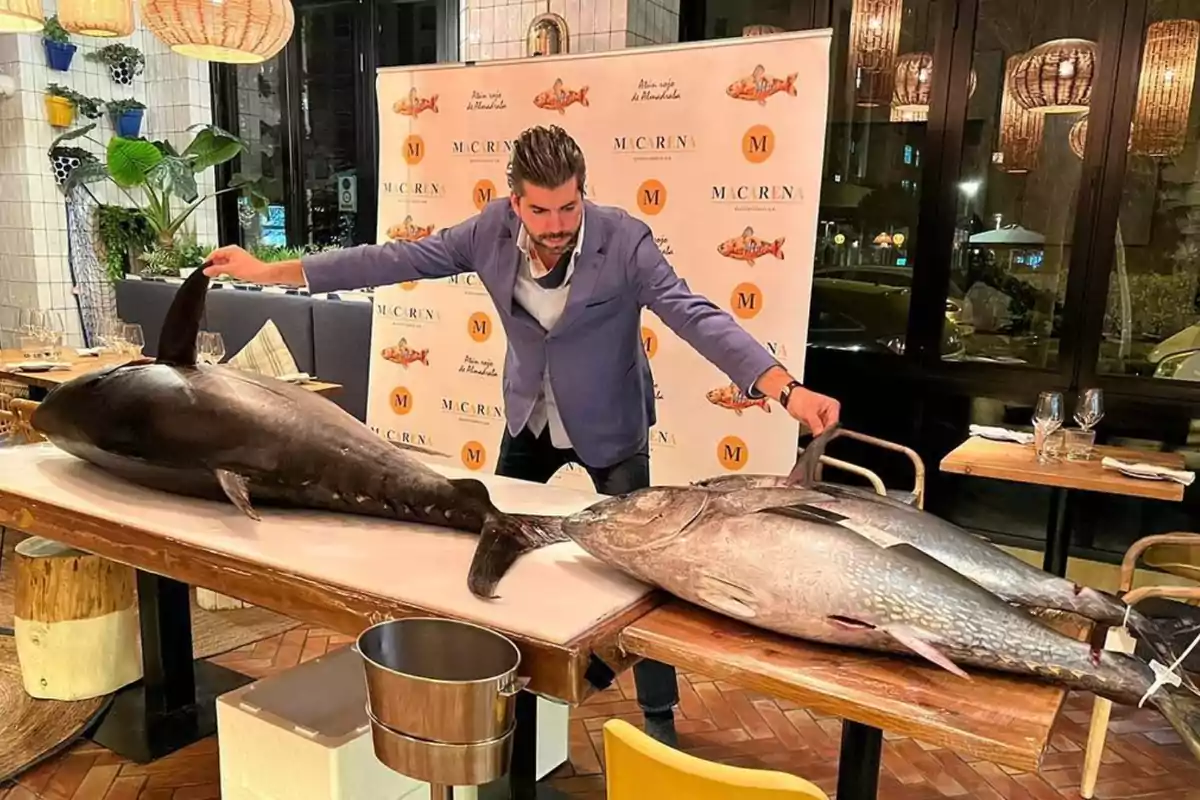 Hombre con chaqueta azul sosteniendo dos grandes atunes sobre una mesa en un restaurante elegante.