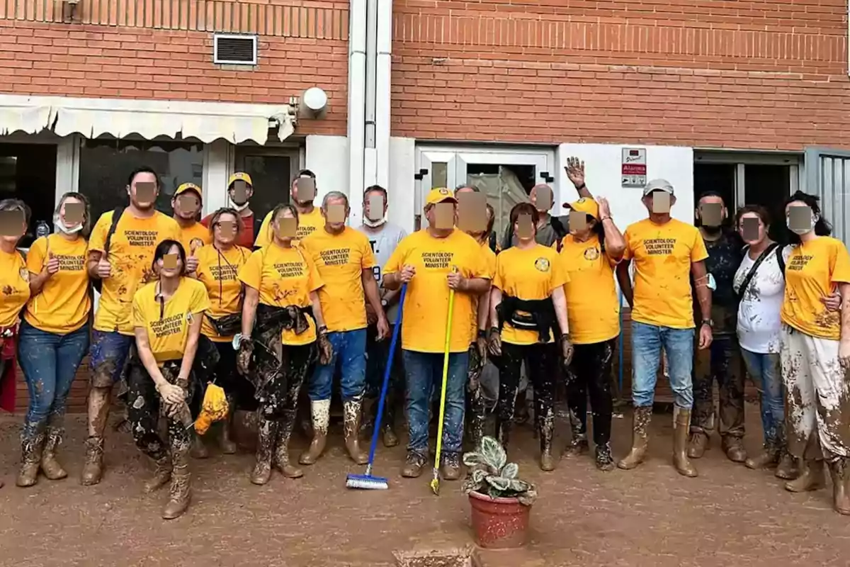 Un grupo de personas con camisetas amarillas y botas embarradas posan frente a un edificio de ladrillo.