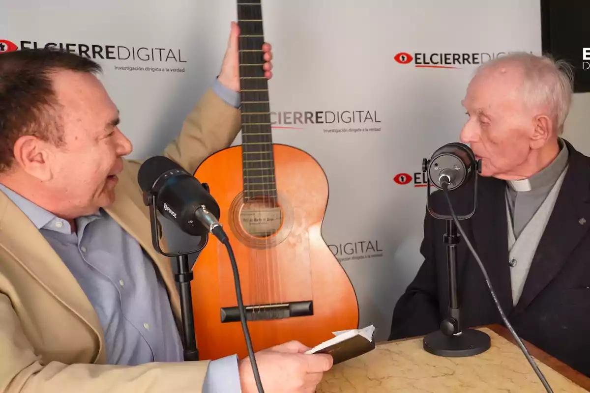 Dos hombres conversan frente a micrófonos en un estudio de grabación mientras uno de ellos sostiene una guitarra.