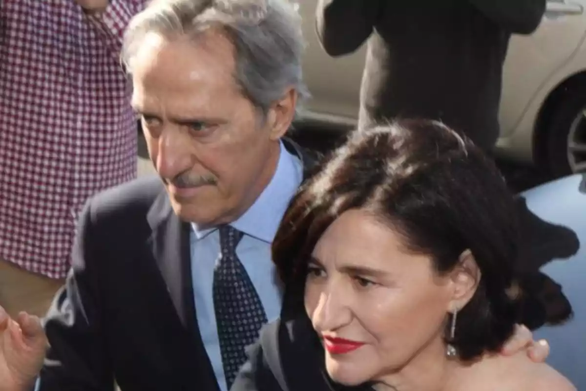 Dos personas caminando juntas, una de ellas con traje oscuro y corbata, la otra con cabello oscuro y labios rojos, rodeadas de otras personas.