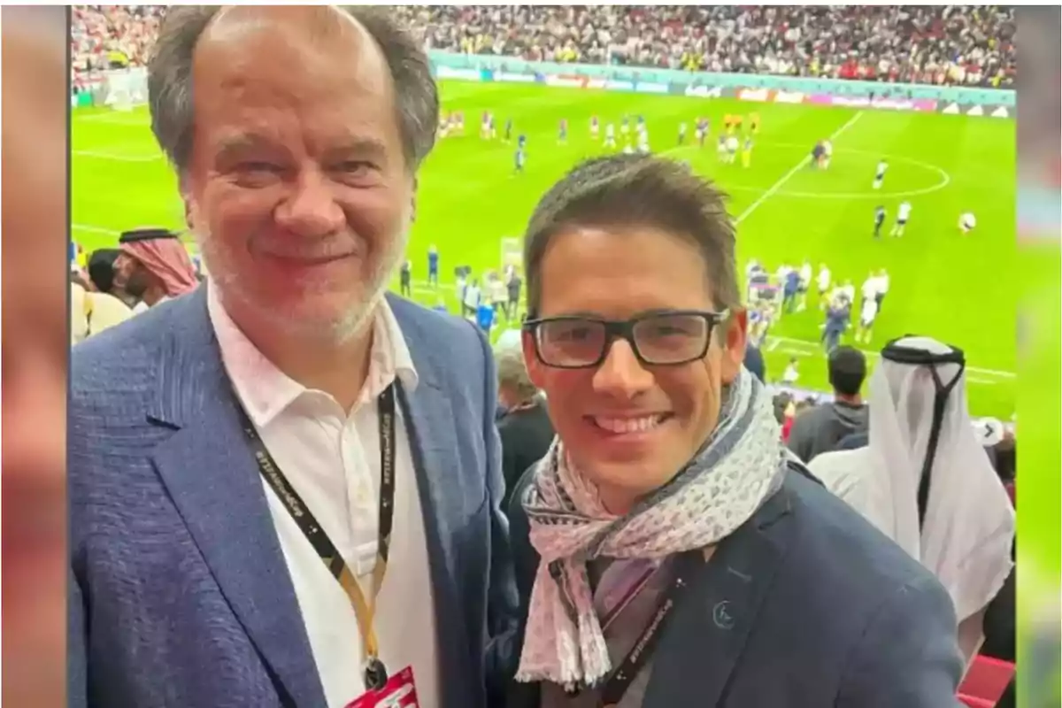 Dos personas sonríen en un estadio de fútbol lleno de espectadores.