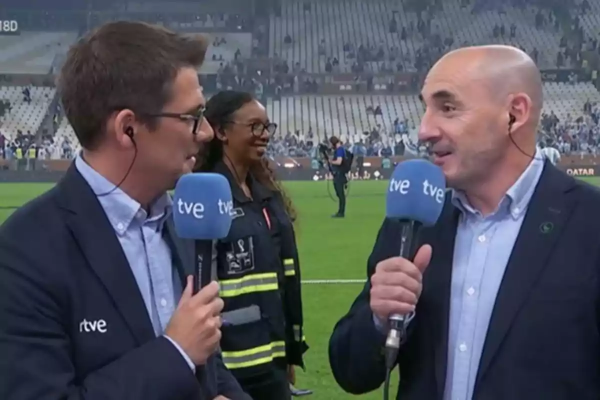 Dos reporteros de TVE con micrófonos en un estadio mientras una persona sonríe en el fondo.