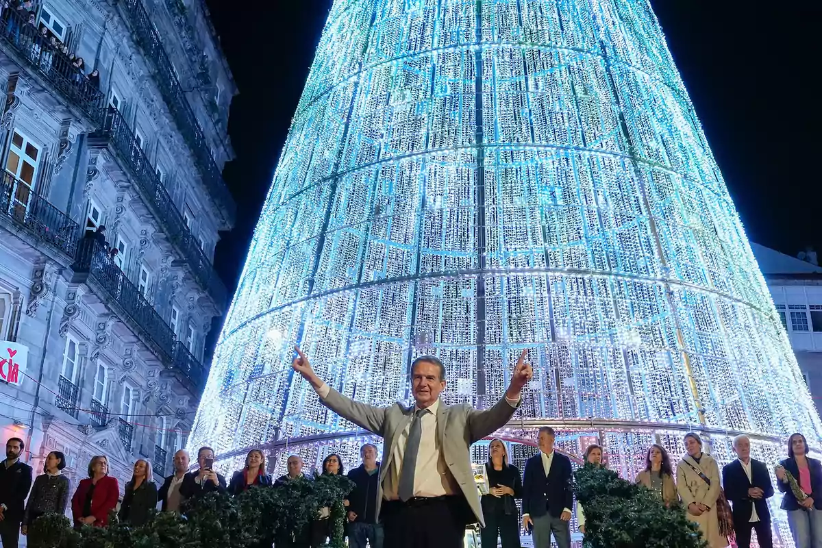 Un hombre con traje y corbata levanta los brazos frente a un gran árbol de Navidad iluminado, rodeado de un grupo de personas y un edificio decorado con luces.