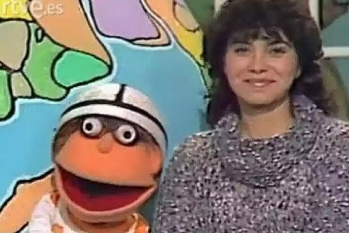 Una mujer sonriente junto a un muñeco de peluche con casco blanco frente a un fondo colorido.