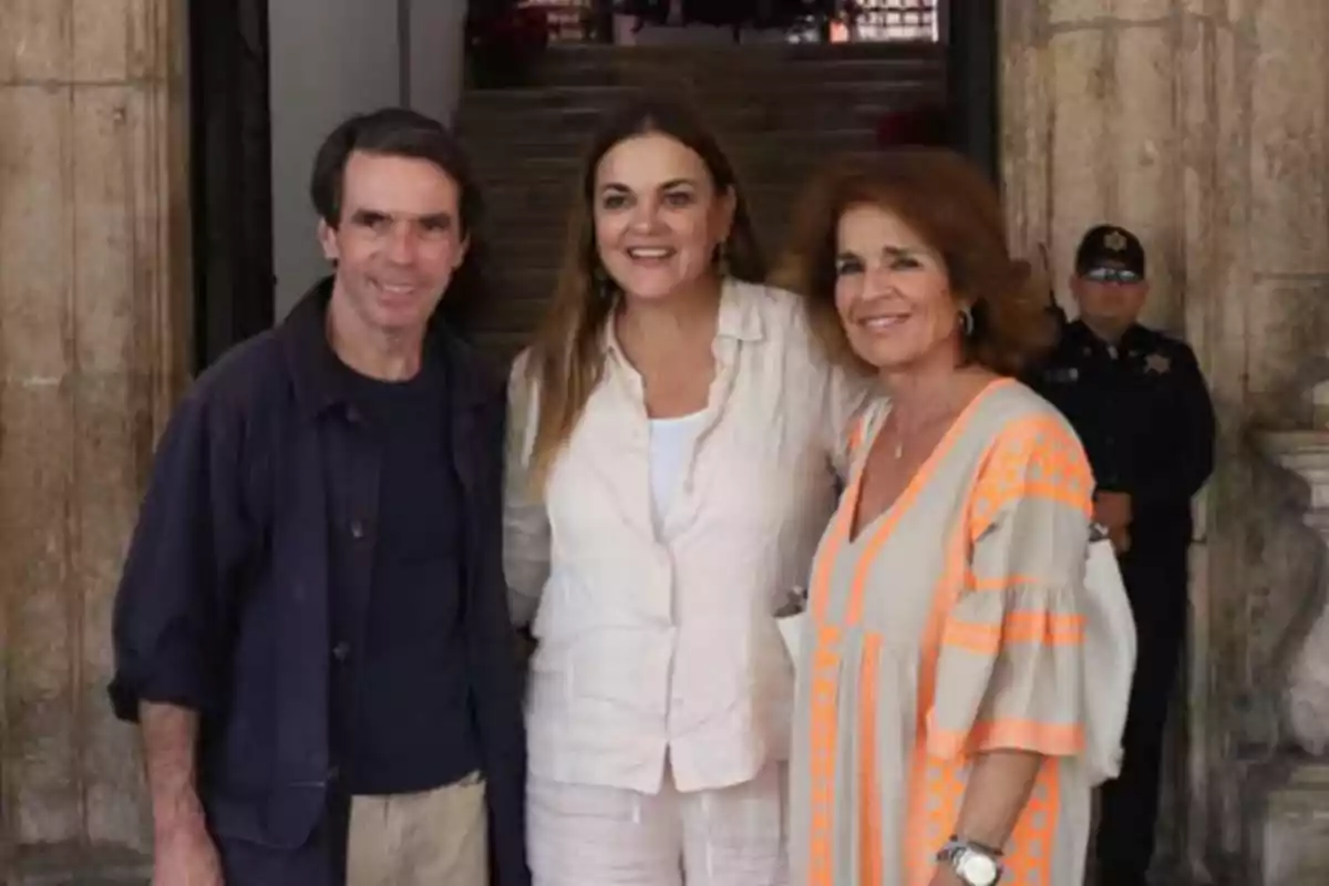 Tres personas posando juntas frente a un edificio con un guardia al fondo.