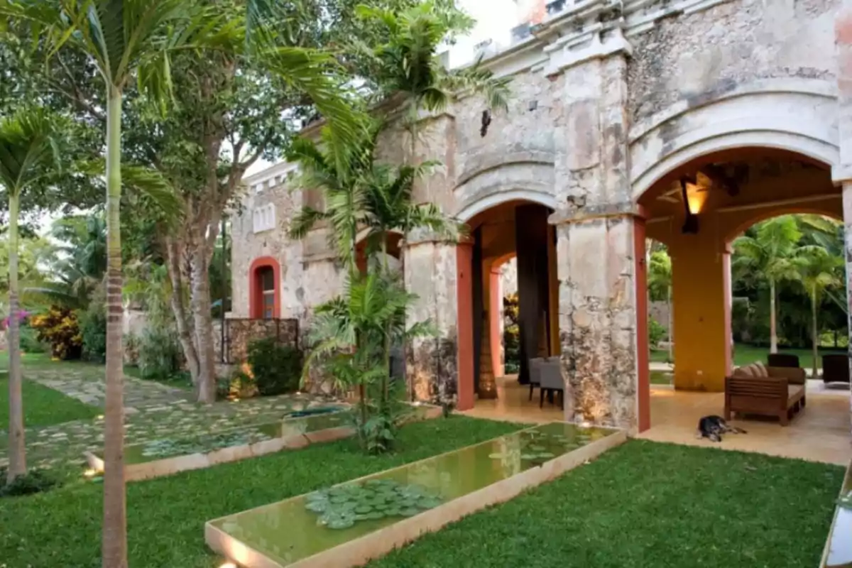 Jardín con palmeras y estanque frente a una antigua construcción de piedra con arcos.