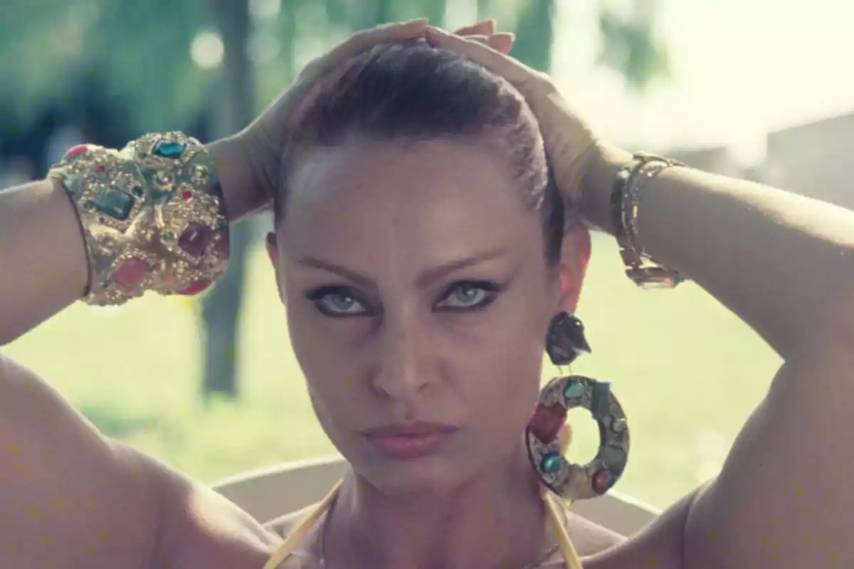 Mujer con cabello recogido y mirada intensa, usando grandes aretes y pulseras doradas con piedras de colores.