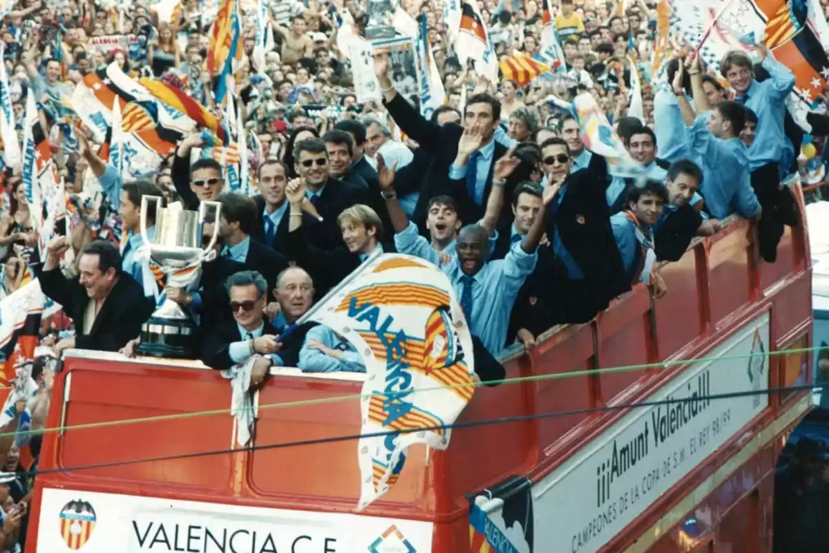 Un grupo de personas celebra en un autobús descapotable con banderas y un trofeo en medio de una multitud.