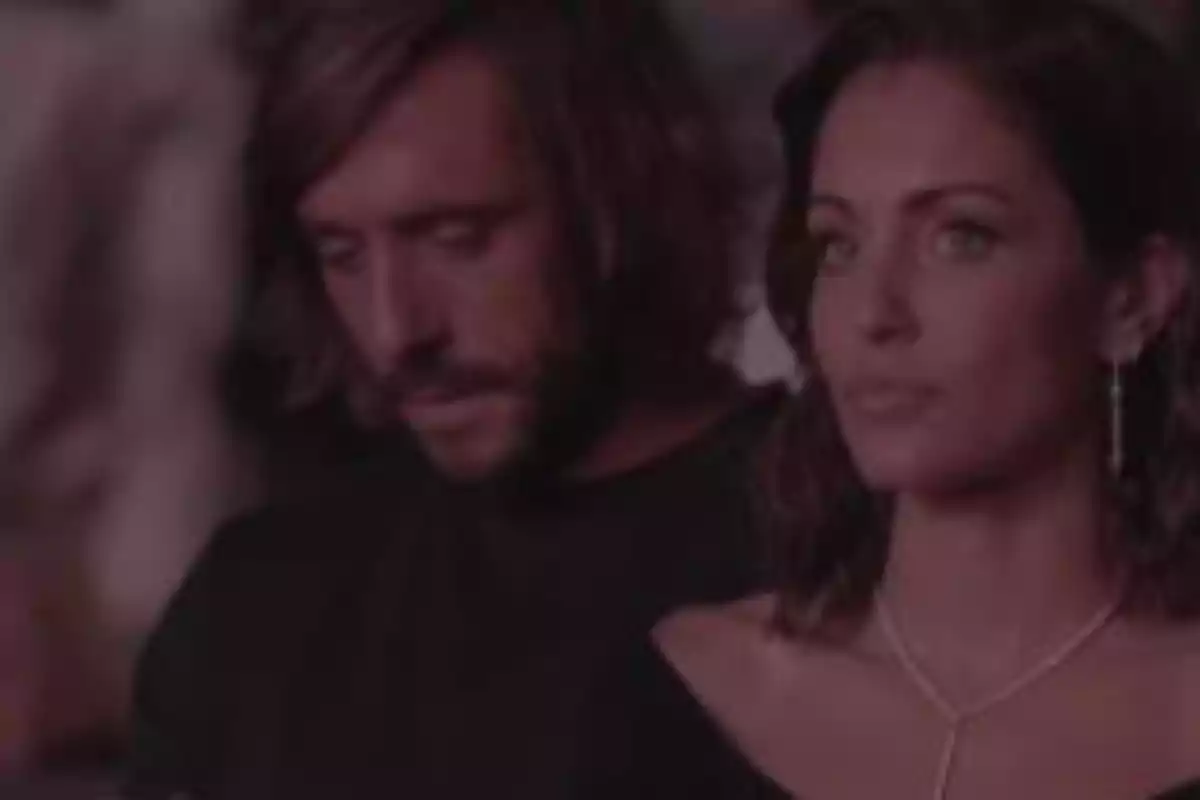 Una pareja sentada, ambos con expresiones serias, en un ambiente con poca luz. Una pareja sentada, ambos con expresiones serias, en un ambiente con poca luz.