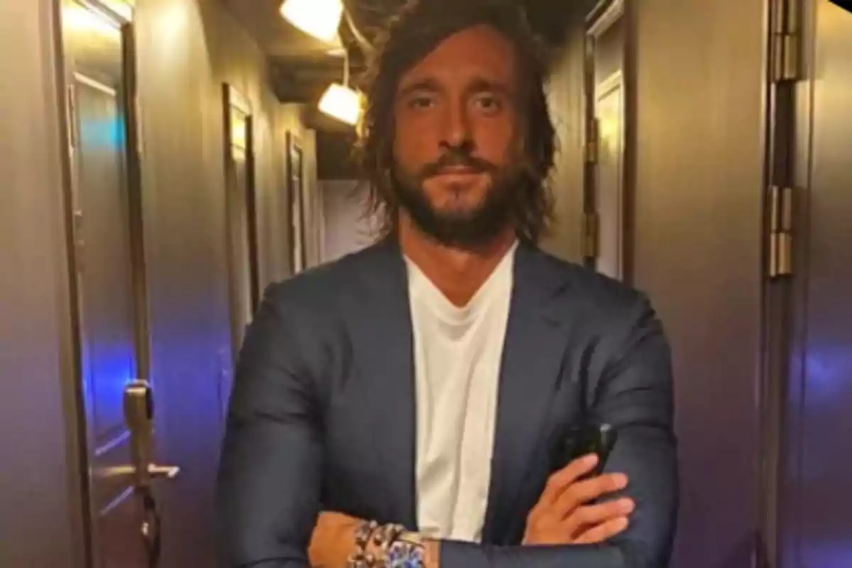 Un hombre con barba y cabello largo viste un traje oscuro y camiseta blanca mientras está de pie en un pasillo iluminado.