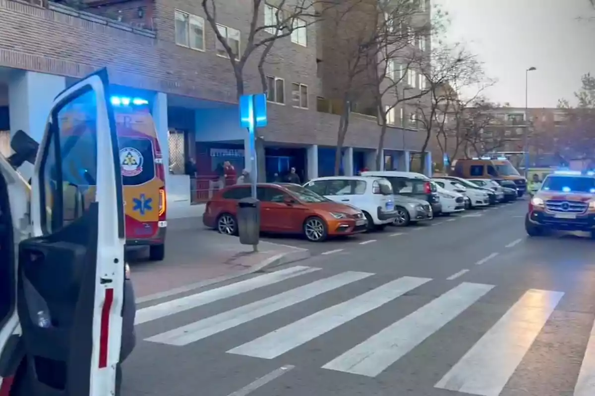 Una calle con varios vehículos estacionados, incluyendo ambulancias con luces encendidas, frente a un edificio residencial.