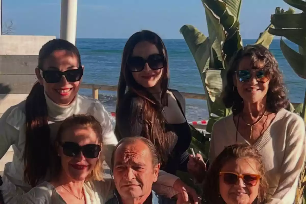 Un grupo de personas posando juntas al aire libre con el mar de fondo y plantas alrededor.