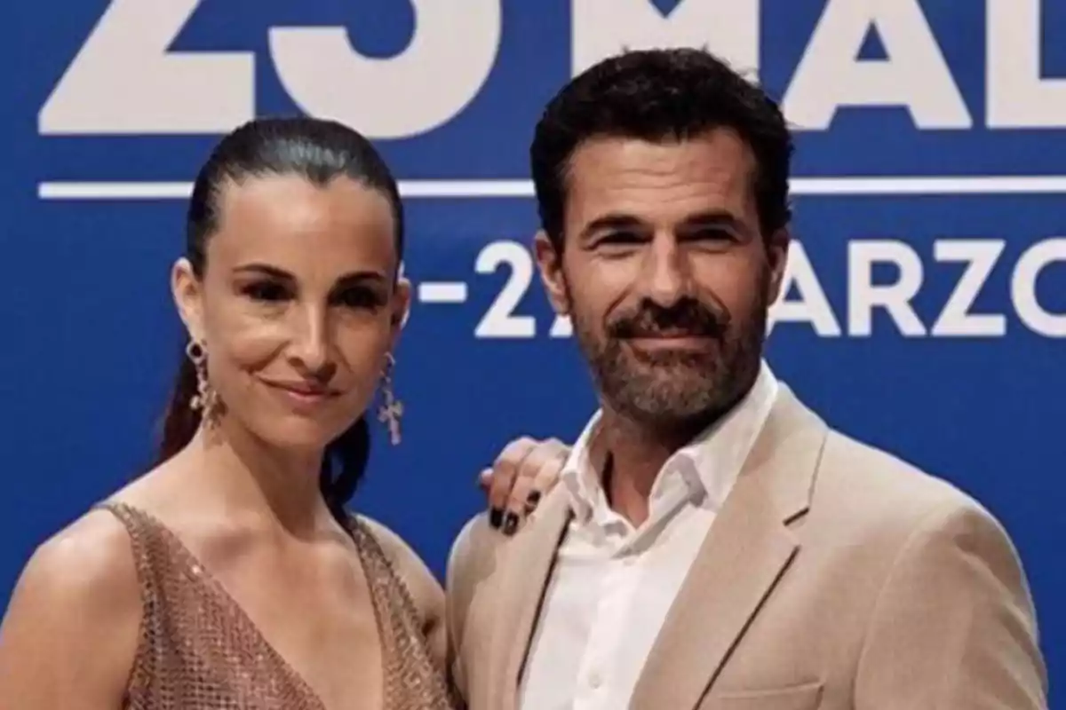 Una pareja posando frente a un fondo azul con texto blanco.