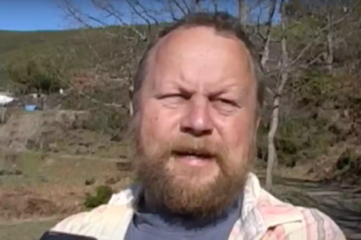 Un hombre con barba en un entorno natural con árboles y colinas al fondo.