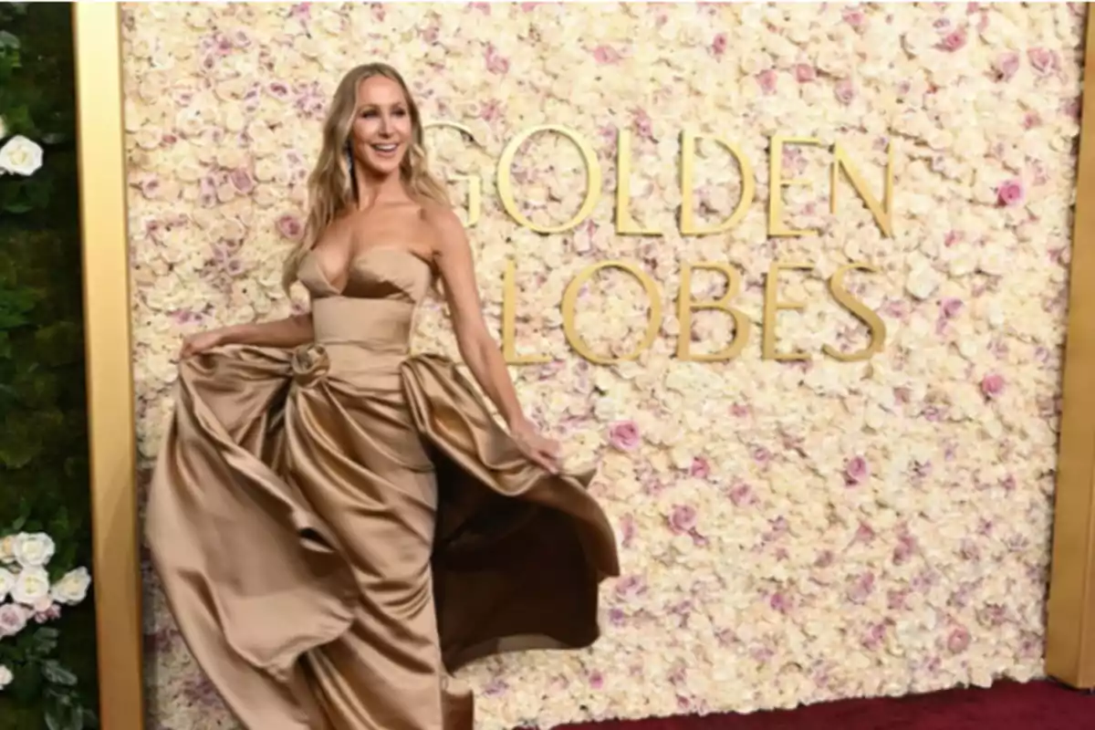 Una persona con un vestido dorado posando frente a un fondo de flores en un evento de alfombra roja.