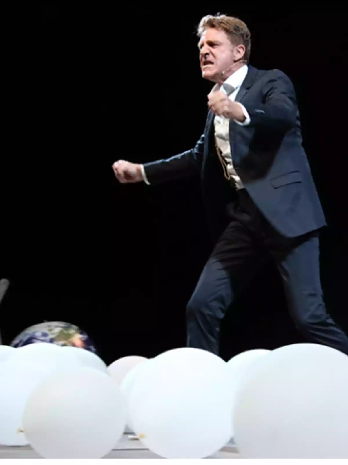 Un hombre en traje oscuro gesticula con intensidad mientras está de pie rodeado de globos blancos sobre un escenario oscuro.