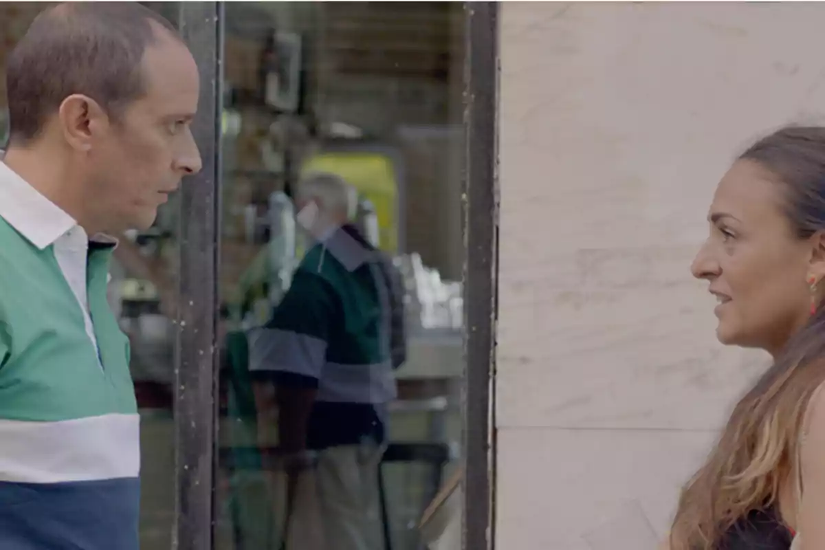 Dos personas conversan frente a un escaparate, reflejando el interior de un local.