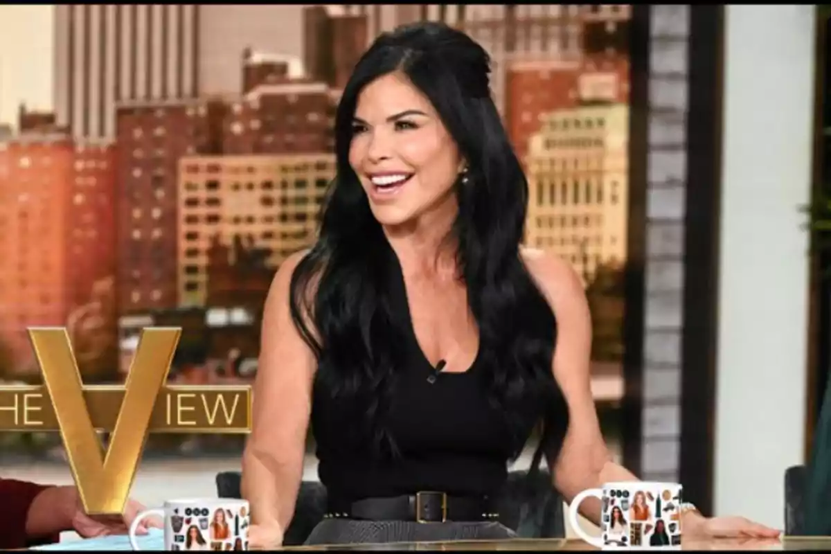 Una mujer de cabello oscuro y largo sonríe mientras está sentada en un set de televisión con tazas decoradas y un fondo urbano.