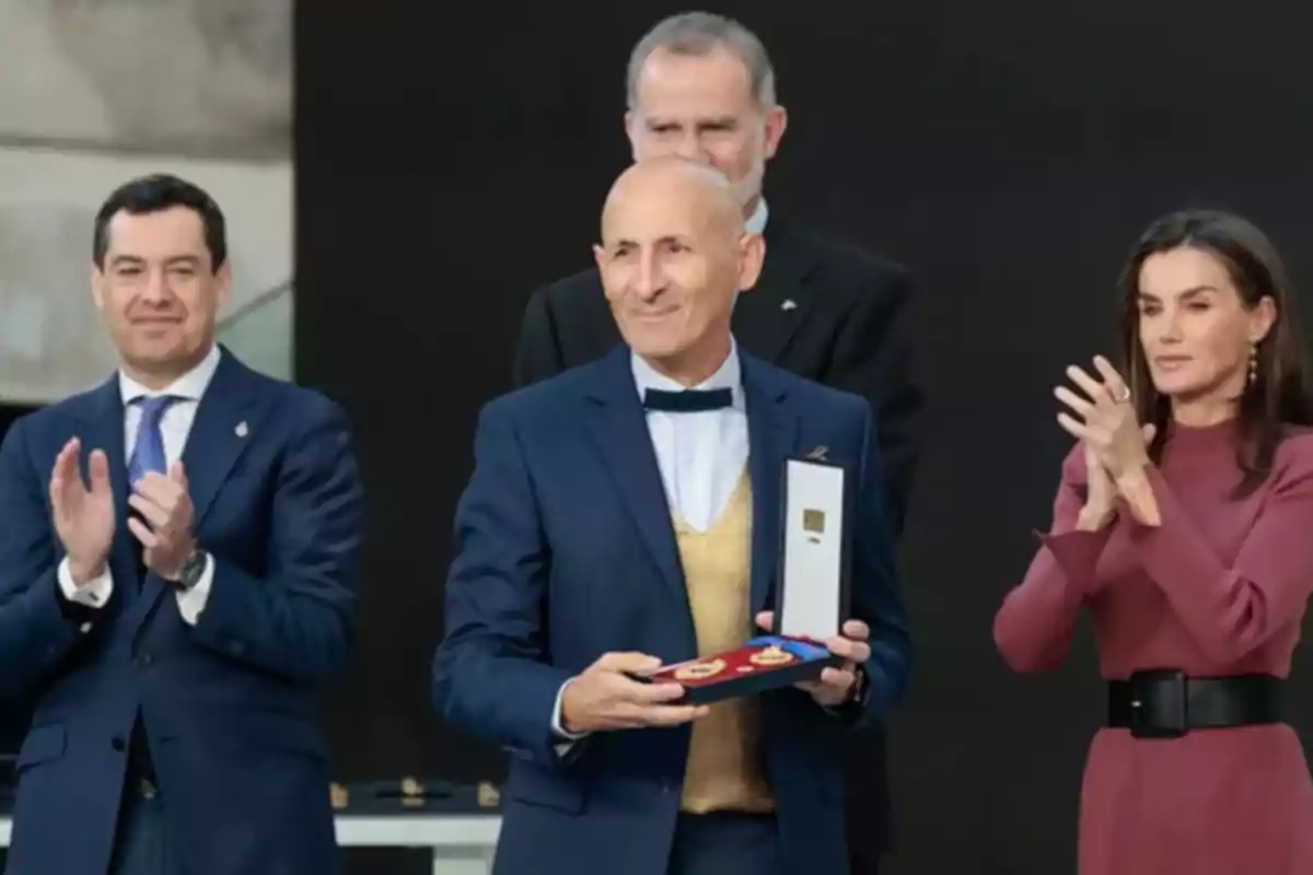 Un hombre sostiene un estuche con medallas mientras otras personas aplauden a su alrededor.