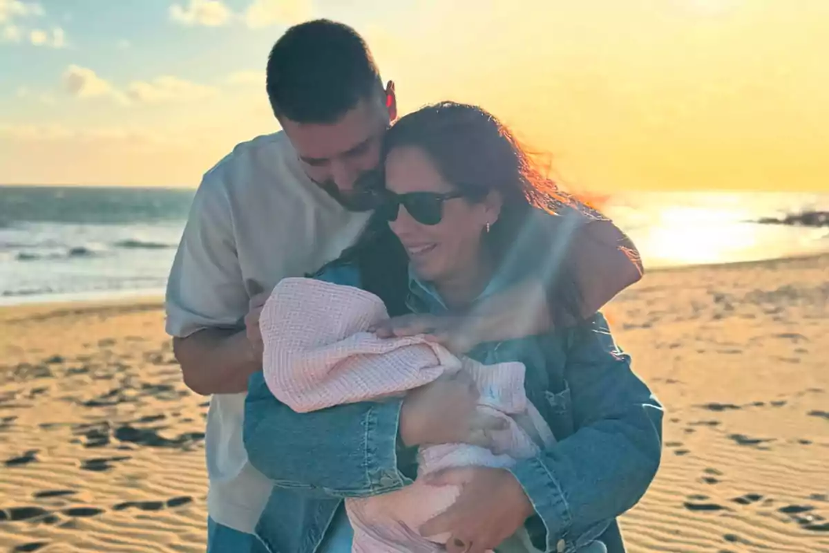 Una pareja sonriente sostiene a un bebé envuelto en una manta en la playa al atardecer.