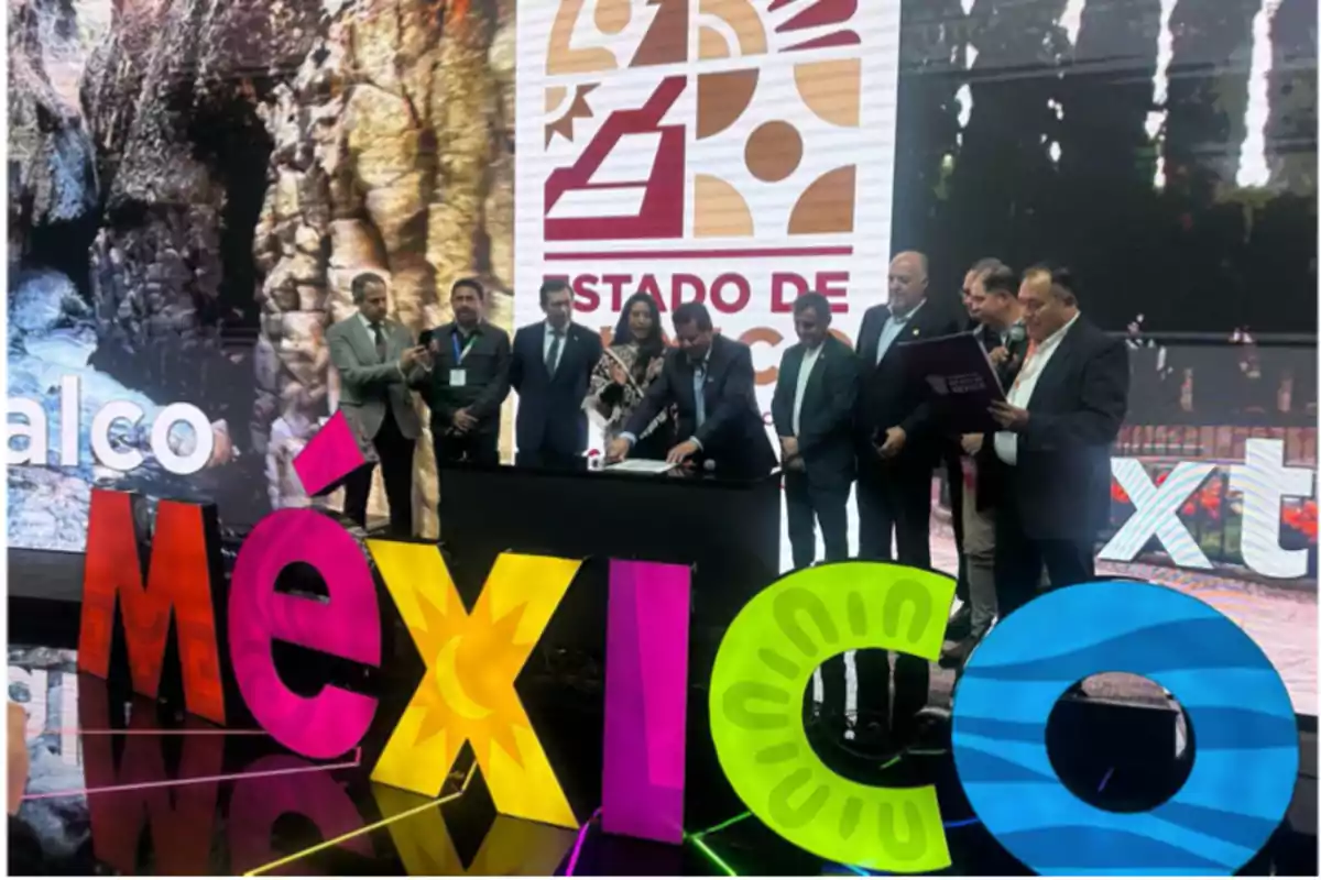 Un grupo de personas está reunido en un evento frente a un letrero colorido que dice "México", con un fondo que muestra el logotipo del Estado de México.