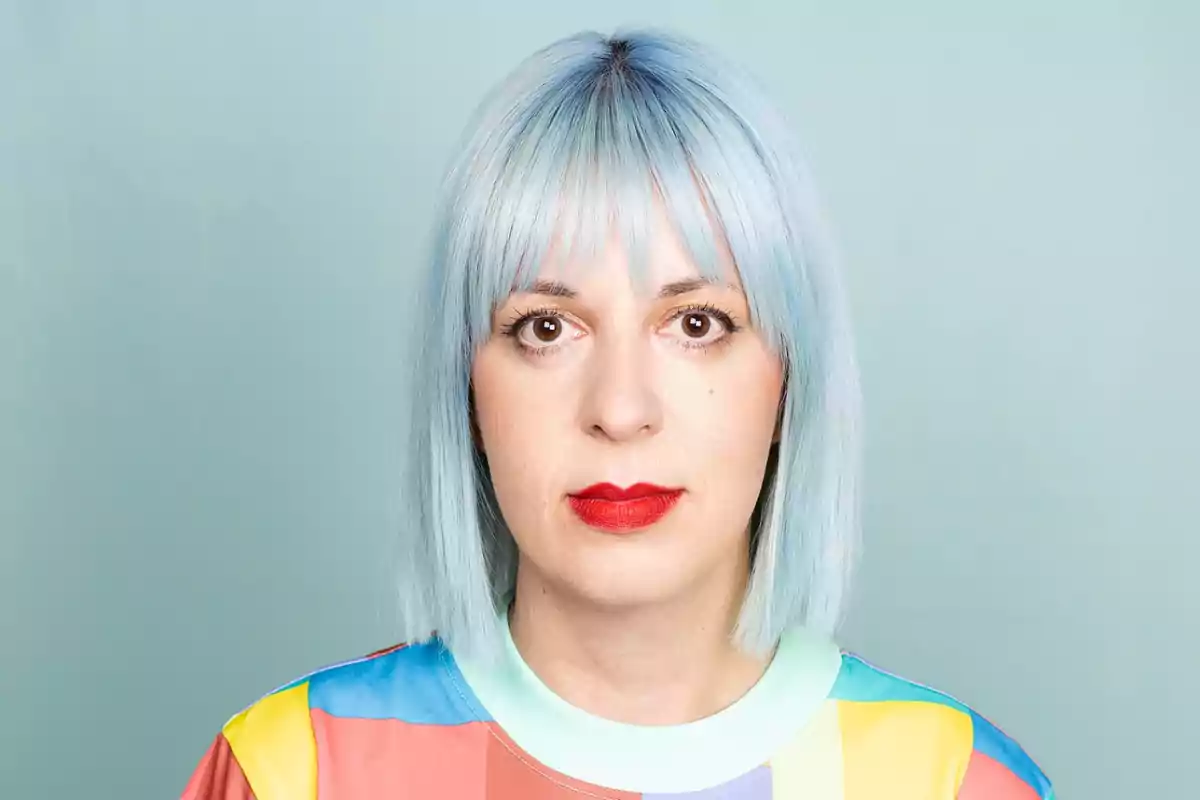 Mujer con cabello azul claro y labios rojos, vistiendo una camiseta colorida, sobre un fondo azul.