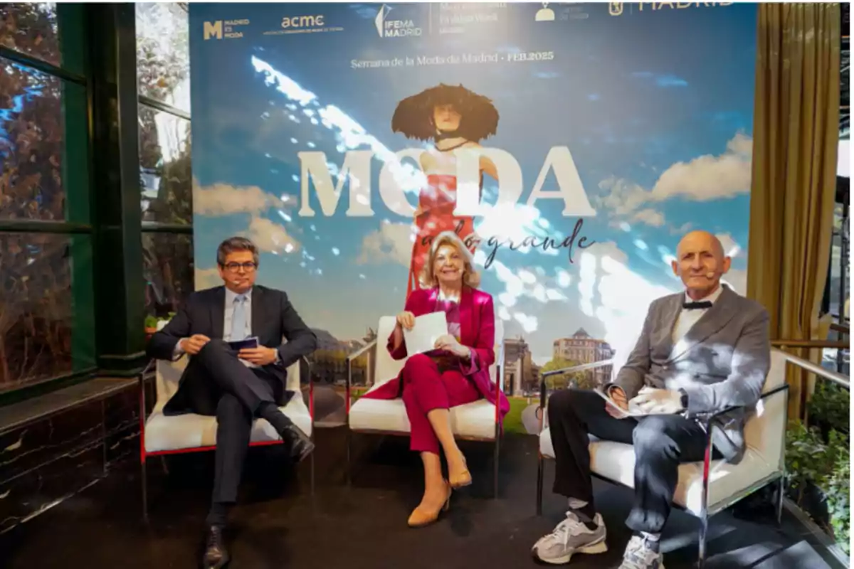 Tres personas sentadas en sillas frente a un cartel de la Semana de la Moda de Madrid.