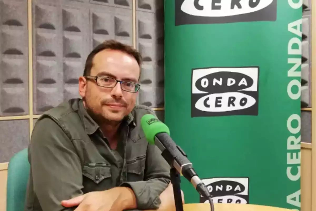 Un hombre con gafas y camisa verde está sentado frente a un micrófono en un estudio de radio con un cartel de Onda Cero en el fondo.