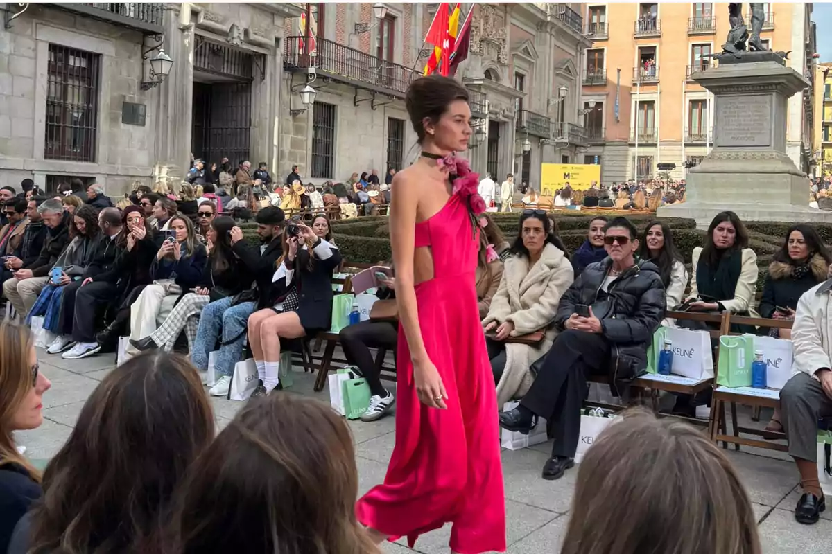 Una modelo camina por una pasarela al aire libre vistiendo un vestido rosa brillante mientras el público observa y toma fotos en un entorno urbano.