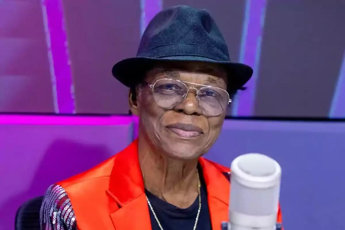 Un hombre mayor con gafas y sombrero negro, vestido con una chaqueta roja, está frente a un micrófono en un estudio iluminado con luces moradas.