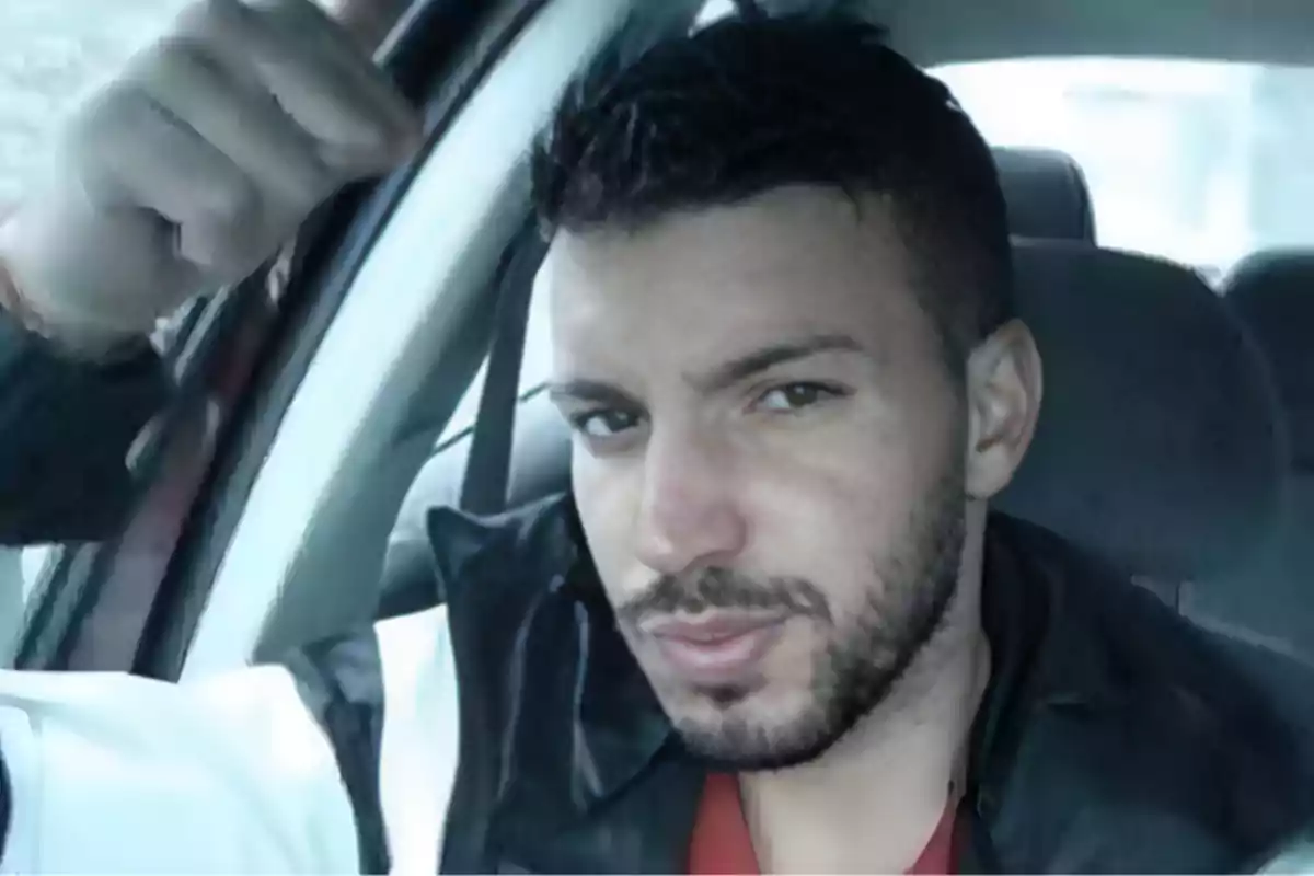 Un hombre con barba y cabello corto está sentado en el interior de un automóvil mirando hacia la cámara.