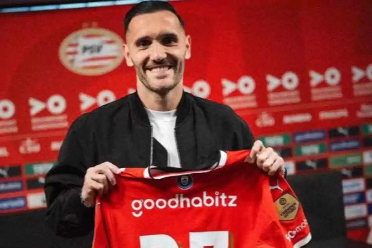 Un hombre sonriente sostiene una camiseta roja de fútbol con un fondo de logotipos y colores rojos.