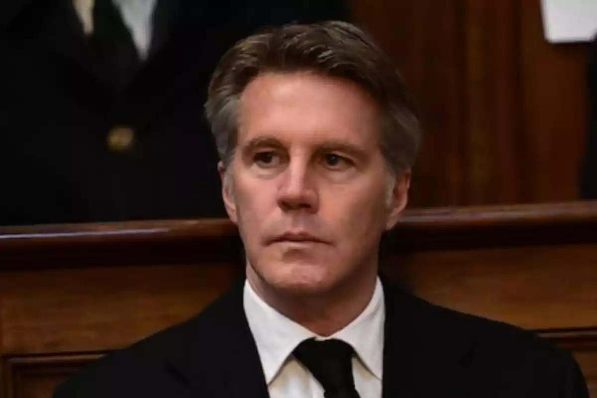 Un hombre de cabello canoso y expresión seria viste traje oscuro y corbata en un entorno formal.