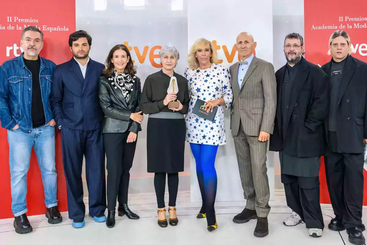 Un grupo de personas posando juntas en un evento de premios, con un fondo que muestra logotipos y texto relacionado con la moda española.