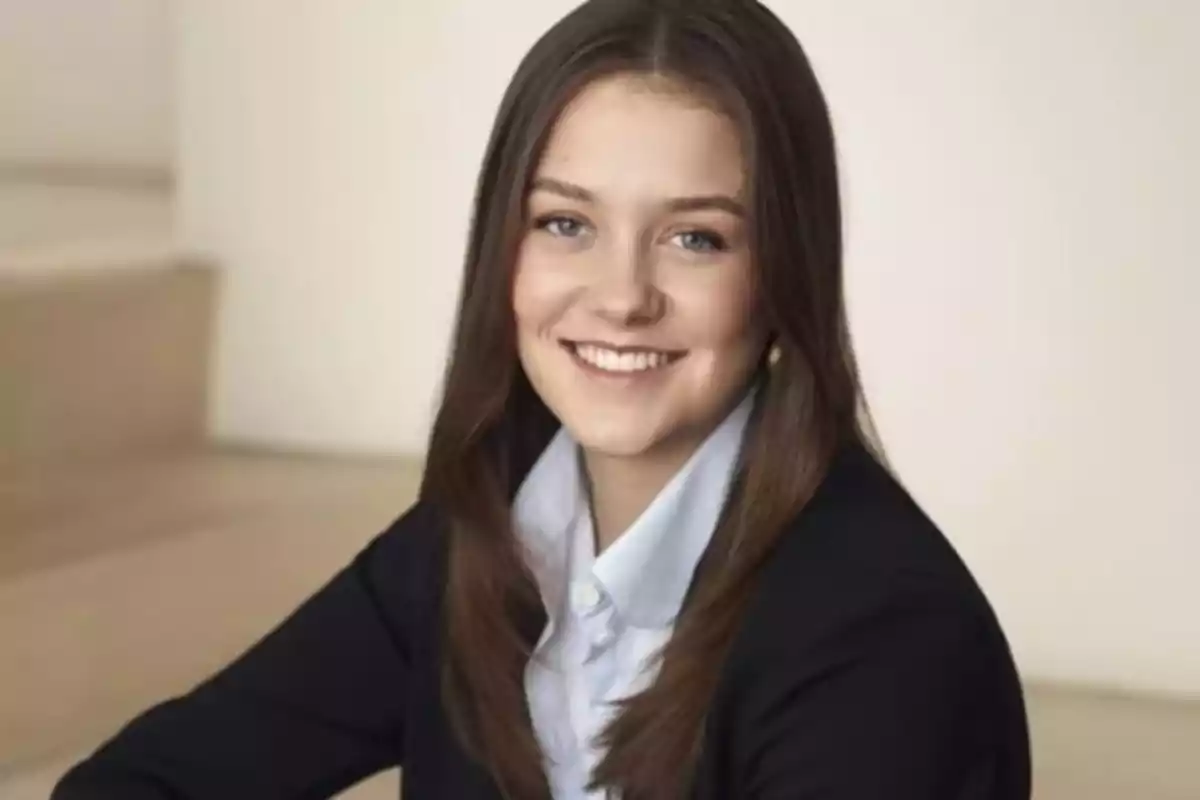 Una joven con cabello castaño y sonrisa, vestida con una chaqueta oscura y camisa clara, en un entorno interior.