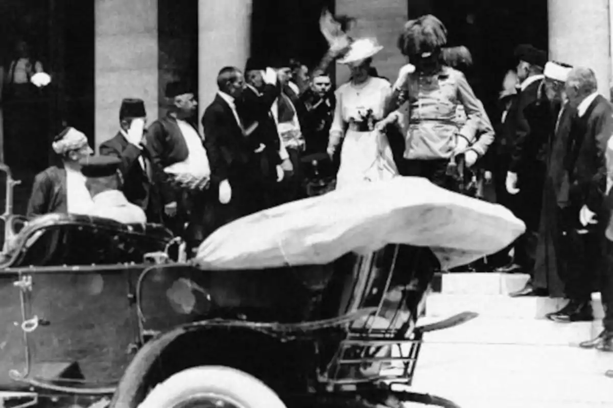 Un grupo de personas vestidas de manera formal se encuentra reunido en la entrada de un edificio mientras un automóvil antiguo está estacionado cerca. Un grupo de personas vestidas de manera formal se encuentra reunido en la entrada de un edificio mientras un automóvil antiguo está estacionado cerca.