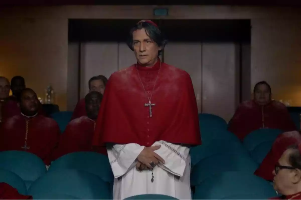 Un hombre vestido con vestimenta eclesiástica roja y blanca está de pie en una sala con varias personas sentadas detrás de él.