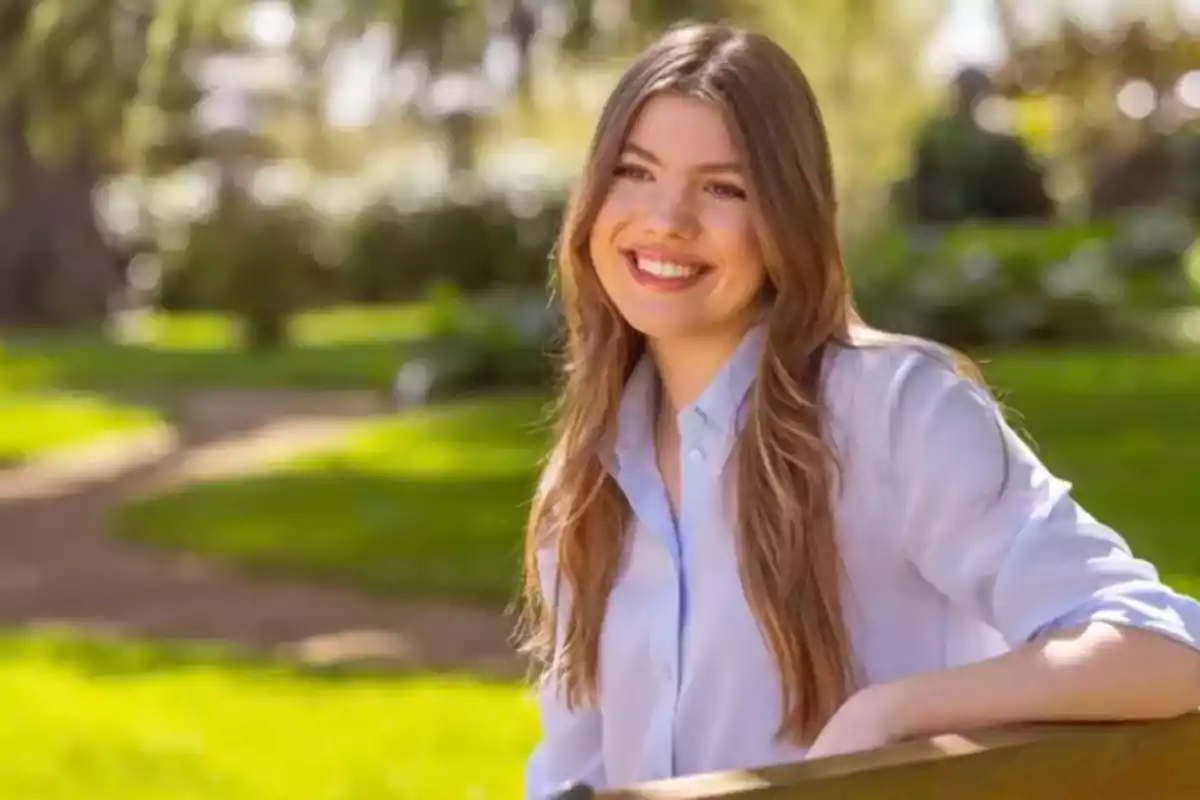 Una mujer sonriente con cabello largo y castaño claro está sentada en un banco de madera en un parque soleado con un fondo de césped verde y árboles.