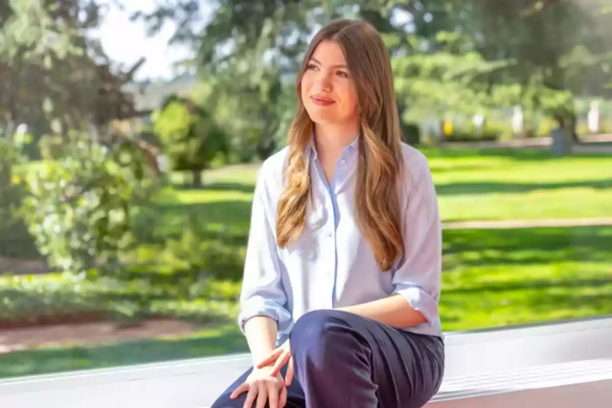Una mujer joven con cabello largo y castaño, vestida con una camisa azul claro y pantalones oscuros, está sentada en un banco frente a un paisaje verde y soleado.